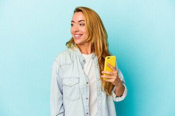 Caucasian woman holding mobile phone isolated on blue background looks aside smiling, cheerful and...