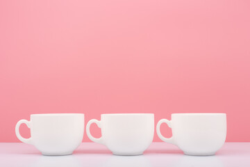 Fototapeta na wymiar Three white shiny ceramic cups in a row on white table against bright pink background with copy space. Creative concept of hot drinks and kitchenware