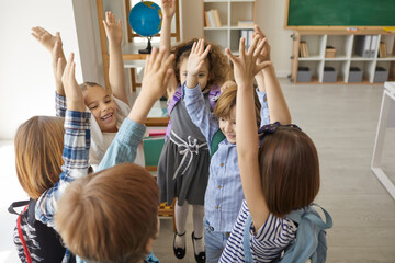 Group of enthusiastic children so glad to be back to school standing in circle with hands up and shouting Hurray. Yay, school's out for summer. Kids having fun in classroom after last class is over