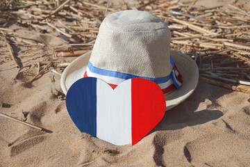 wooden heart and straw hat with ribbon with colors of flag of France blue, white and red in golden sand on beach