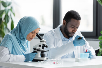 Muslim scientist looking through microscope near blurred african american colleague working in laboratory