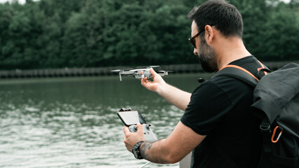 A man with a backpack flying a drone with a phone controller next to the lake. General focus.