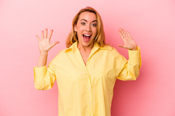 Caucasian blonde woman isolated on pink background receiving a pleasant surprise, excited and raising hands.