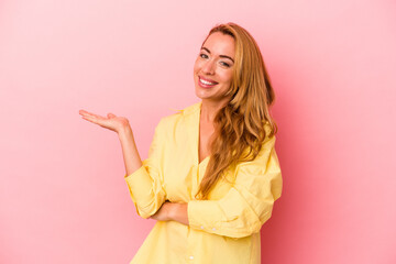 Caucasian blonde woman isolated on pink background showing a copy space on a palm and holding another hand on waist.