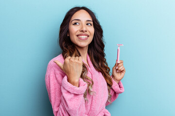 Young mexican woman wearing a bathrobe holding a knife isolated on blue background points with thumb finger away, laughing and carefree.