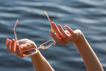 Horizontal close-up conceptual photography with a hands of young white woman, holding sunglasses against blue water surface during relaxing on the beach at summer daytime