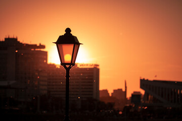 Landscape photography with a vintage lamp post on the foreground and modern buildings silhouettes on the background during orange summer sunset