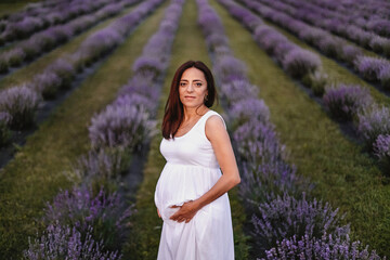 Front view of smiled brunette caucasian pregnant woman dressed in white dress, touching belly in lavender field. Future mother walking in park at summer day. Expecting twins. Concept of maternity.