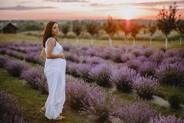 Fototapeta na wymiar View in full height of happy woman expecting newborn child, standing at field with violet lavender and dreaming on sunset. Future mother looking to side, dressed in white outfit. Concept of pregnancy
