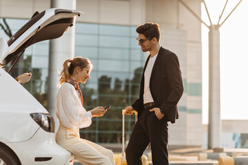 Joyful woman in beige outfit and brunette man in black suit talk near white car. Blonde lady holds passport. Handsome guy poses with yellow suitcase and smiles.