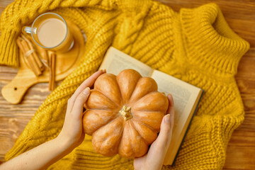 Ripe pumpkin in women's hands over knitted sweater. Aromatic latte with cinnamon sticks on wooden tray. Orange sweater, open romantic book and pumpkin on wooden background. Autumn composition.