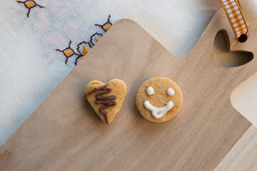 Top view of two cookies located on a wooden kitchen board. One cookie is heart-shaped and the other is smiley-shaped.