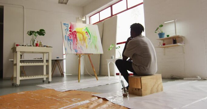 Rear view of thoughtful african american male artist sitting looking at his painting at art studio
