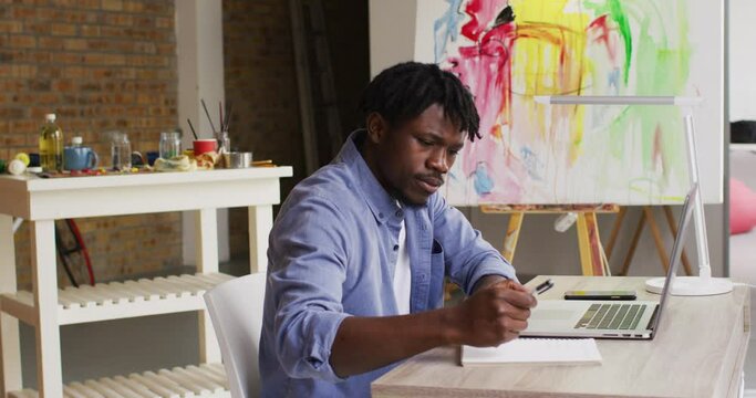 African american male artist taking notes while sitting on his desk at art studio