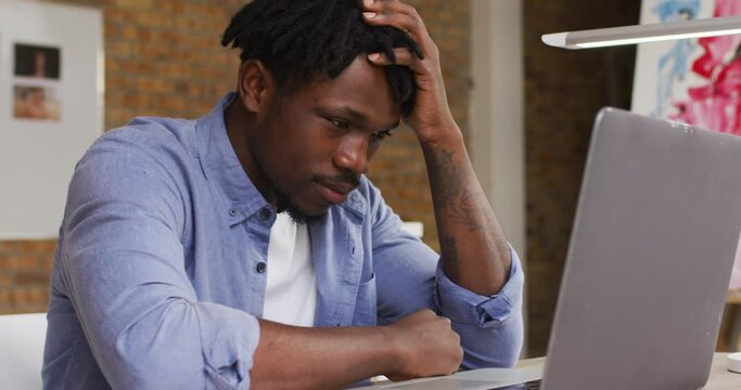 Stressed african american male artist looking at his laptop while sitting on his desk at art studio