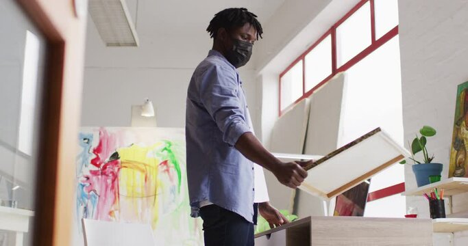 African american male artist wearing face mask holding a painting and using laptop at art studio