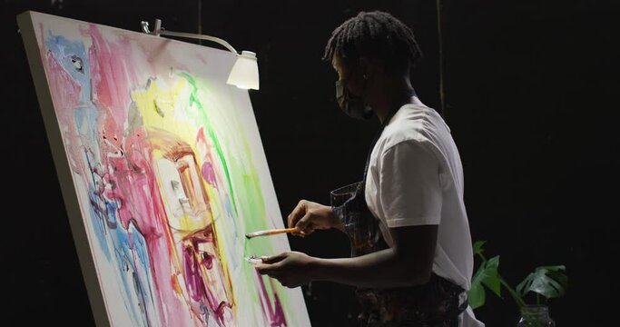 African american male artist wearing face mask painting on canvas at art studio