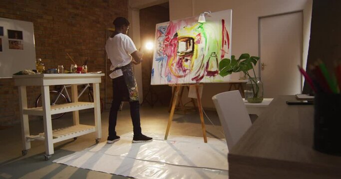 African american male artist wearing apron painting with palette knife on canvas at art studio