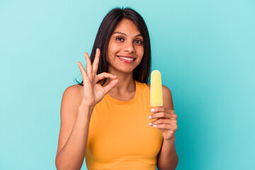 Young latin woman holding ice cream isolated on blue background cheerful and confident showing ok gesture.