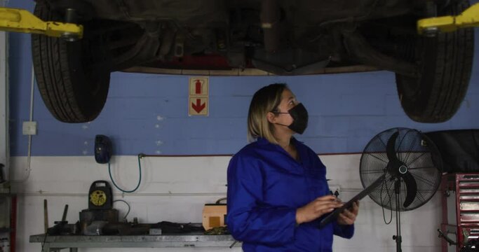 Female mechanic wearing face mask holding clipboard and inspecting the car at a car service station