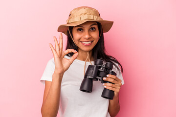 Young latin woman holding binoculars isolated on pink background cheerful and confident showing ok gesture.