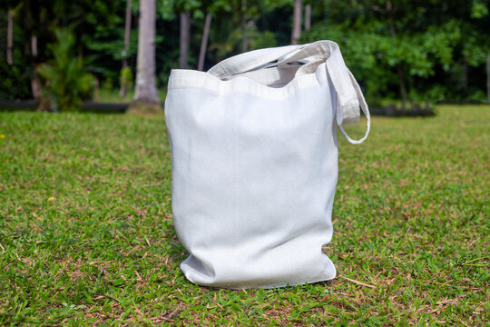 Canvas tote bag on sunny green lawn. Simple tote bag mockup. Summer landscape with green lawn and forest. Beach wear mock-up. White bag front view. Textile print template