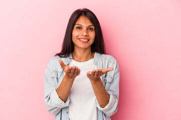 Young latin woman isolated on pink background holding something with palms, offering to camera.