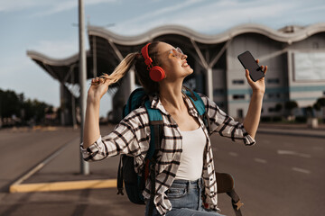 Fototapeta na wymiar Charming blonde girl in sunglasses, red headphones listens to music. Pretty female tourist holds phone and backpack. Attractive woman in plaid shirt poses near airport.