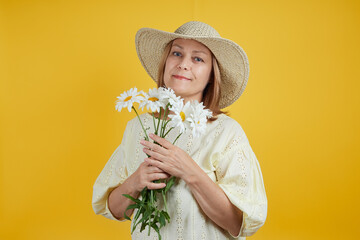 Beautiful mature woman in a straw hat with chamomile in her hands smiling on a bright yellow background. The concept of summer sales, summer vacation, travel with copy space