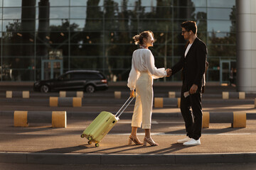 Happy blonde woman in white blouse, beige pants holds yellow suitcase and greets with man in black suit near airport.
