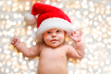 Obraz na płótnie Canvas Beautiful little child is celebrating Christmas. New Year's holidays. A child in a Christmas costume. Childhood and people concept - happy Newborn baby in Santa hat over holidays lights background