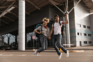 Young active man in white tee, jeans holds passport. Couple of travelers jumps with backpacks. Happy woman and guy in sunglasses smiles.