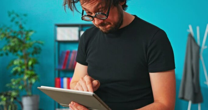 Funny, crazy, strange boy sitting on table with tablet in hand, clicks on screen, shocked look, quickly looks at camera opens face and gestures surprise and astonishment, rolls eyes, blinks few times