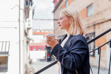 Fototapeta na wymiar Happy beautiful young woman in dress and jacket walks around the city and drinks coffee
