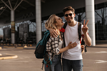 Portrait of happy brunette man in sunglasses showing peace sign and stylish blonde woman near airport. Charming curly girl hugs boyfriend and holds backpack.