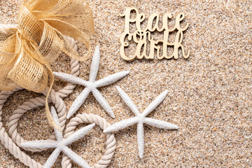 beach or tropical theme holiday greeting with white starfish, nautical rope, mesh net bow and Peace on Earth on beach sand all in natural tones