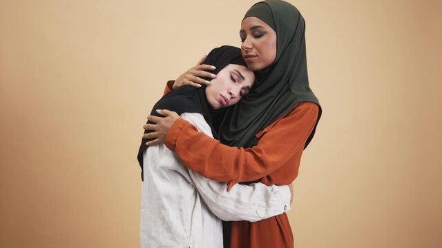 An emotional arabian woman wearing national hijab is hugging her friend giving her support standing in the studio isolated over beige background