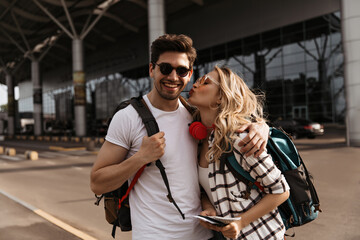 Curly woman kisses her boyfriend near airport. Attractive girl in plaid shirt and brunette man in white tee poses in good mood and holds backpacks.