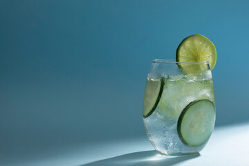 Vaso con agua mineral y hielos, con rodajas de pepino y limón sobre un fondo de color azul