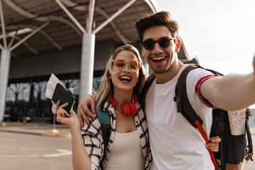 Blonde girl in sunglasses and red headphones holds passport and tickets. Man in white tee takes...