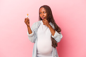 Young african american pregnant woman holding pacifier isolated on pink background looking sideways with doubtful and skeptical expression.