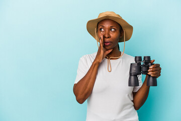 Young african american woman holding binoculars isolated on blue background is saying a secret hot...