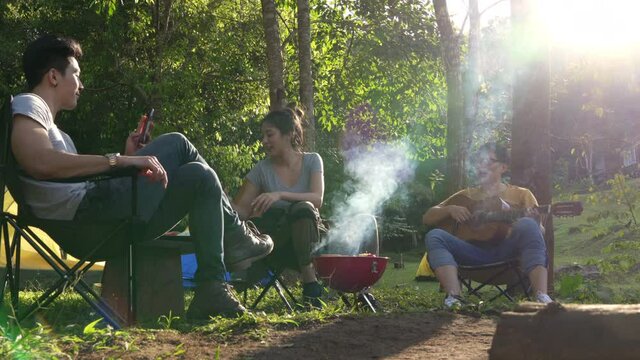 Group of Asian people friends sitting on outdoor chair eating barbecue grill for dinner and singing together. Man and woman friendship relax and enjoy outdoor lifestyle activity camping on vacation.