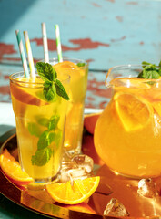 Glasses of lemonade with lemons, oranges and mint leaves on blue wooden background. Summer cold drinks under direct sun shines.