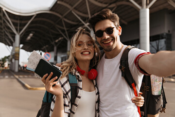 Pretty blonde woman in sunglasses, plaid shirt holds plane tickets and passport. Brunette man hugs girlfriend and takes selfie near airport. Portrait of travelers.