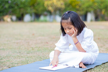 Little adorable Asian kid lying down on mat writing on notebook, Concept of outdoor education, A girl doing her homework, Kid like reading book