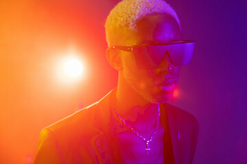 a guy  blond hair looking into the camera. He has dark glasses on his face. Against the background of bright orange light. He is wearing a black jacket and a silver ornament around his neck - 445196548