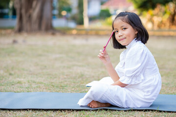 Little adorable Asian kid lying down on mat writing on notebook, Concept of outdoor education, A girl doing her homework, Kid like reading book