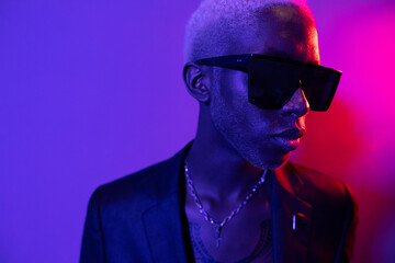 Portrait photo of a dark-skinned guy with a beard. Dressed in a black jacket and sunglasses. He has metal jewelry on his neck. Purple and pink light