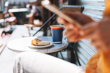 Paper cup with coffee and piece of cheesecake on table in outdoor cafe on background of blurred young woman with the mobile phon
